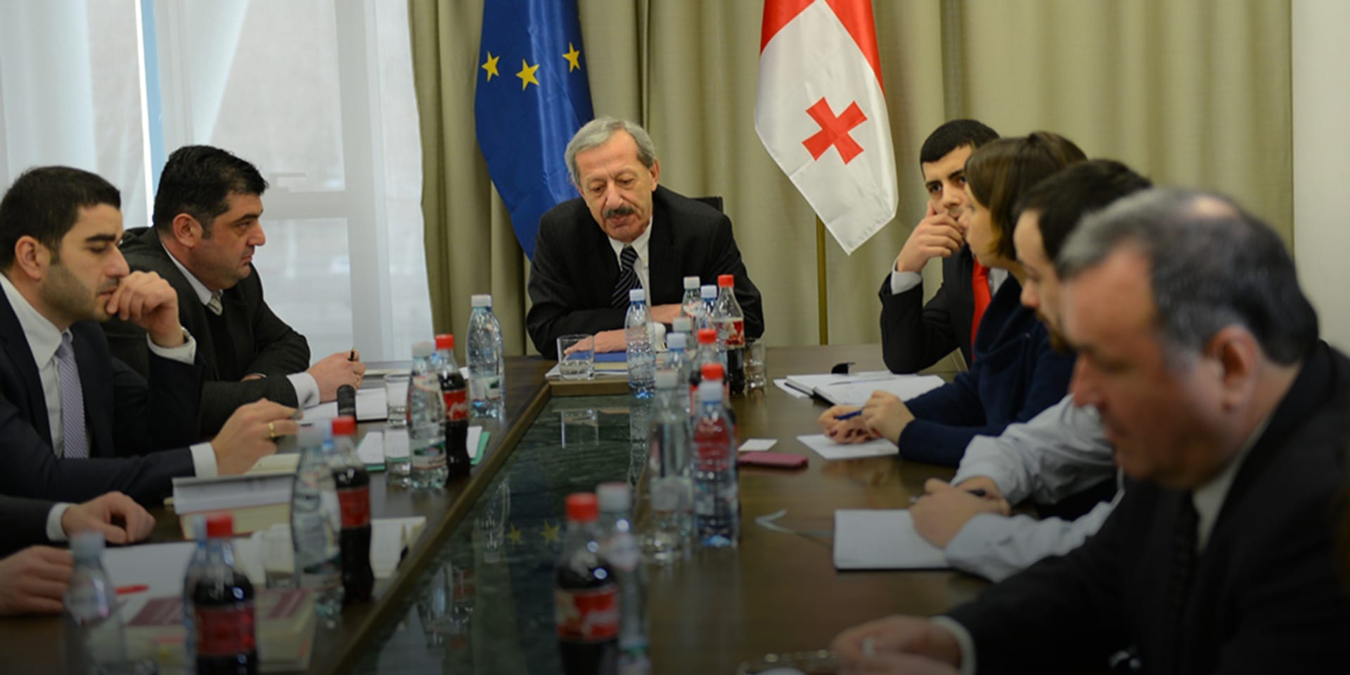 Image of a group of businesspeople having a meeting at a large table with the flags of the EU and Georgia in the background.
