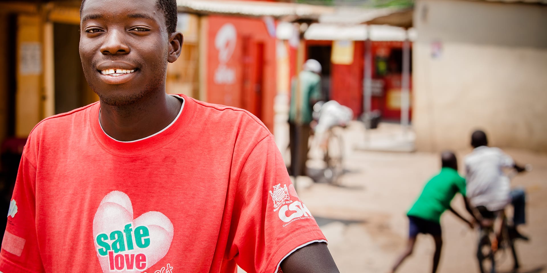Image of a smiling young man on a busy street wearing a red t-shirt that says 