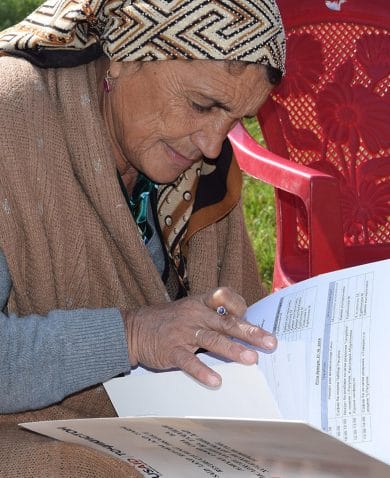 An older woman kneeling beside red plastic chairs on the grass while filling out USAID documents.