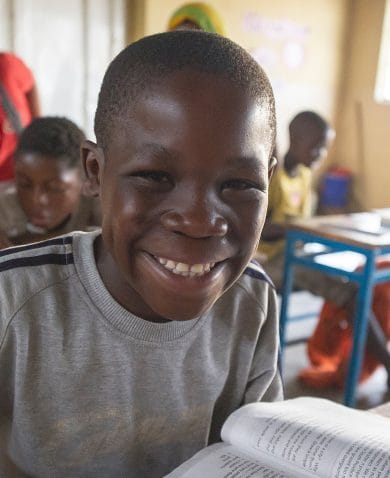 A smiling boy sitting at a school desk holding a pen and reading a book.