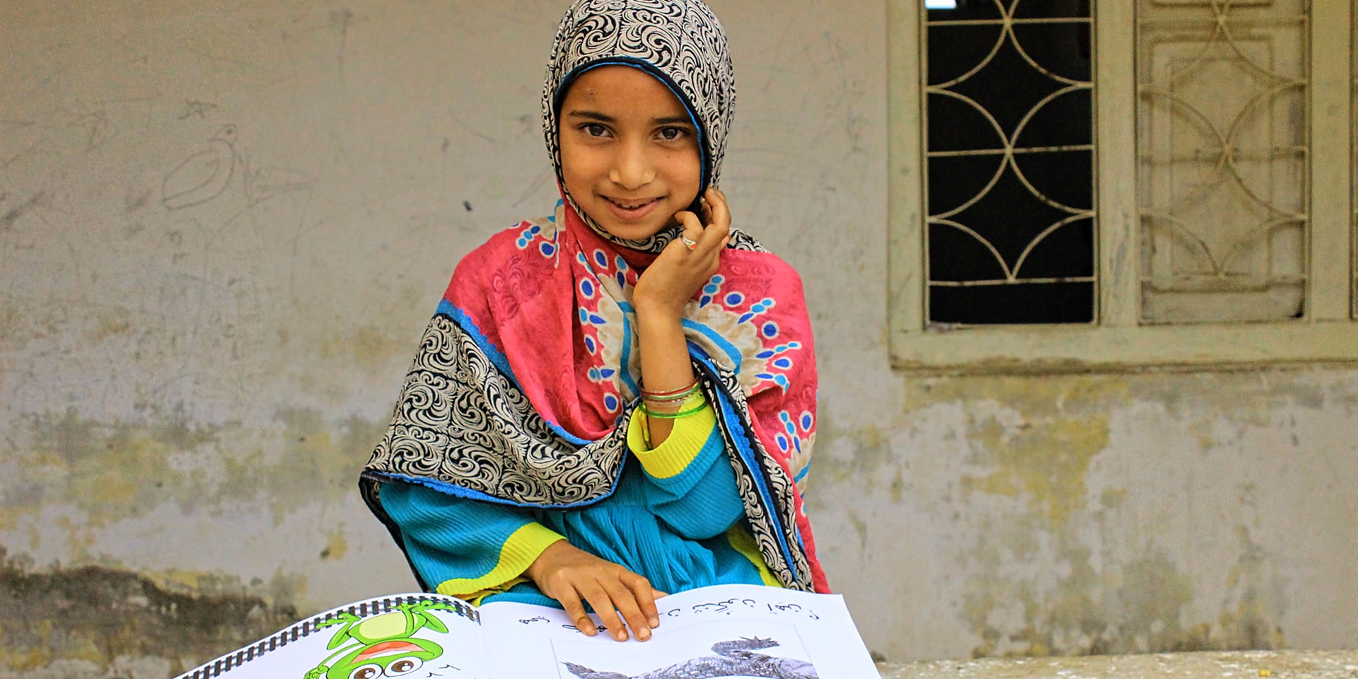 A smiling girl sitting in front of a home and reading from a children's book.