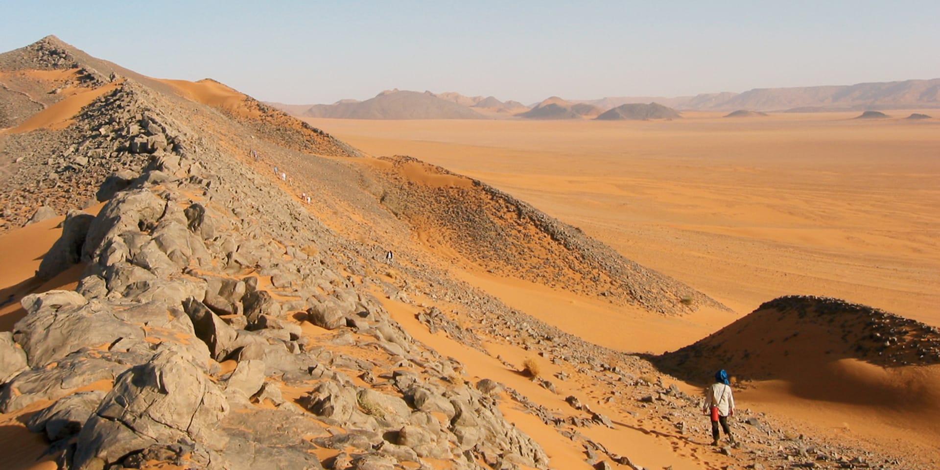 Image of a rocky hill in the desert with a person walking along its base. In the distance are several other large hills.