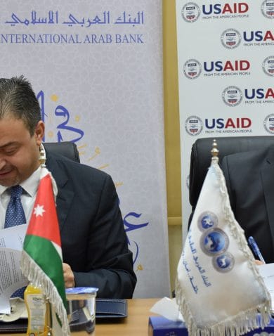 Two smiling men signing documents at a table with United States and Jordan Flags at its center.