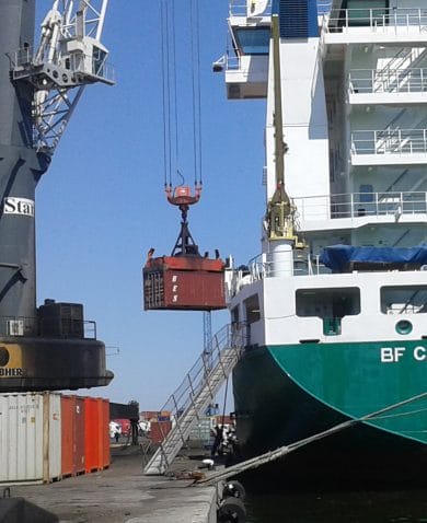Image of a dock with a shipping container being loaded by a crane onto a large green and white boat.