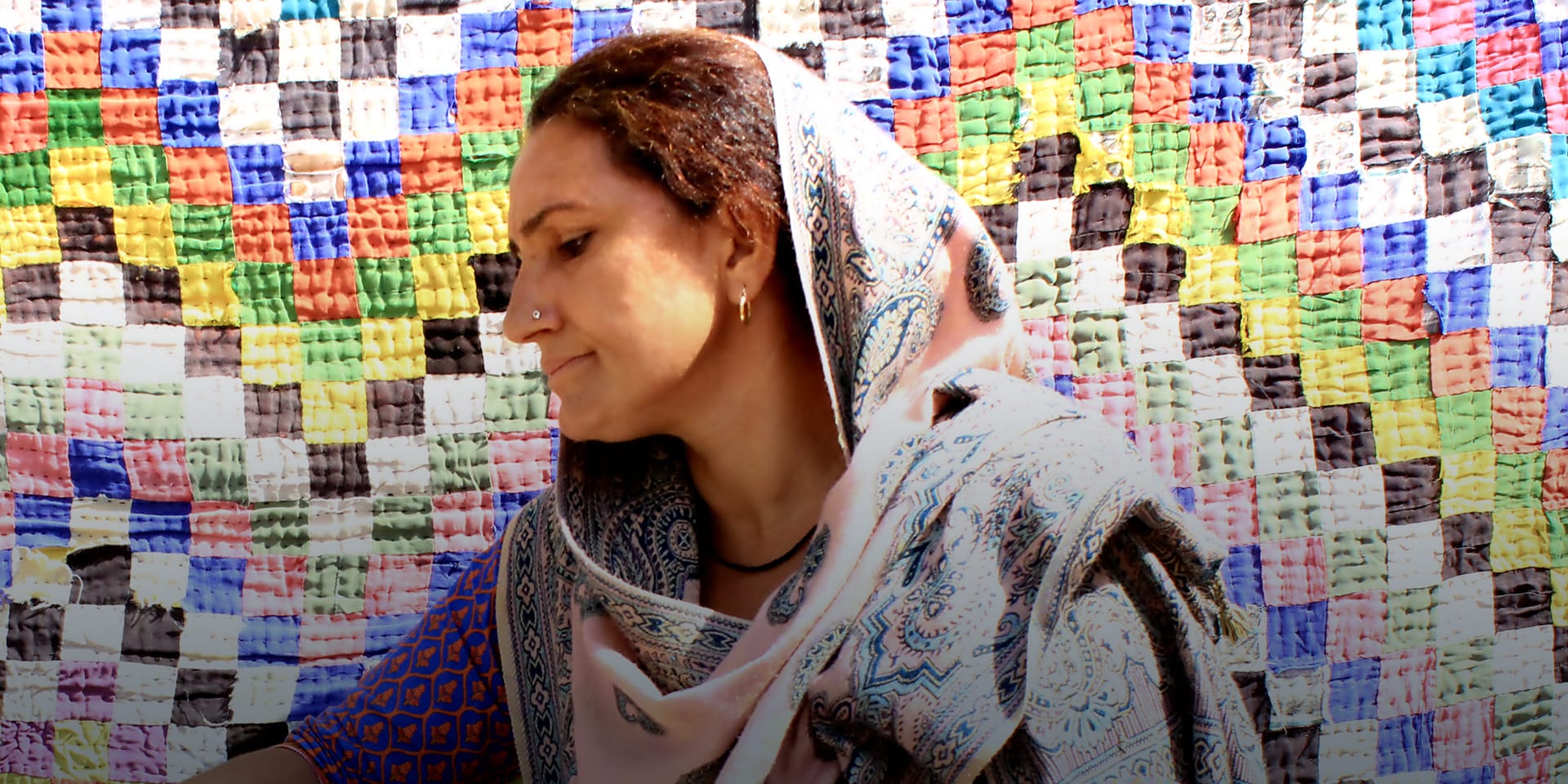 A woman looking off-camera and sitting in front of a large quilt composed of multi-colored squares.