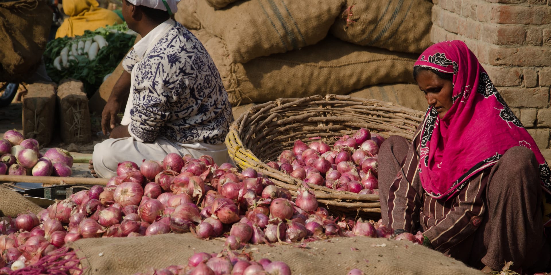 A large pile of onions with a woman sitting beside them, individually peeling them, placing them in a large basket beside her.