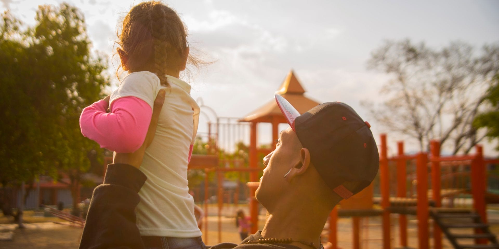 Image of a man in a baseball cap holding up a small girl in a playground.