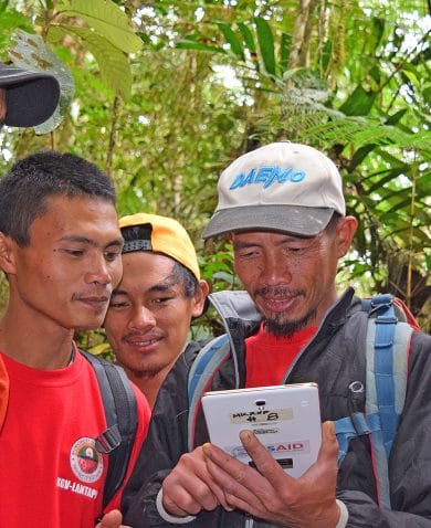 A man showing a tablet he is holding to four other men standing alongside him. They are in a green forest.