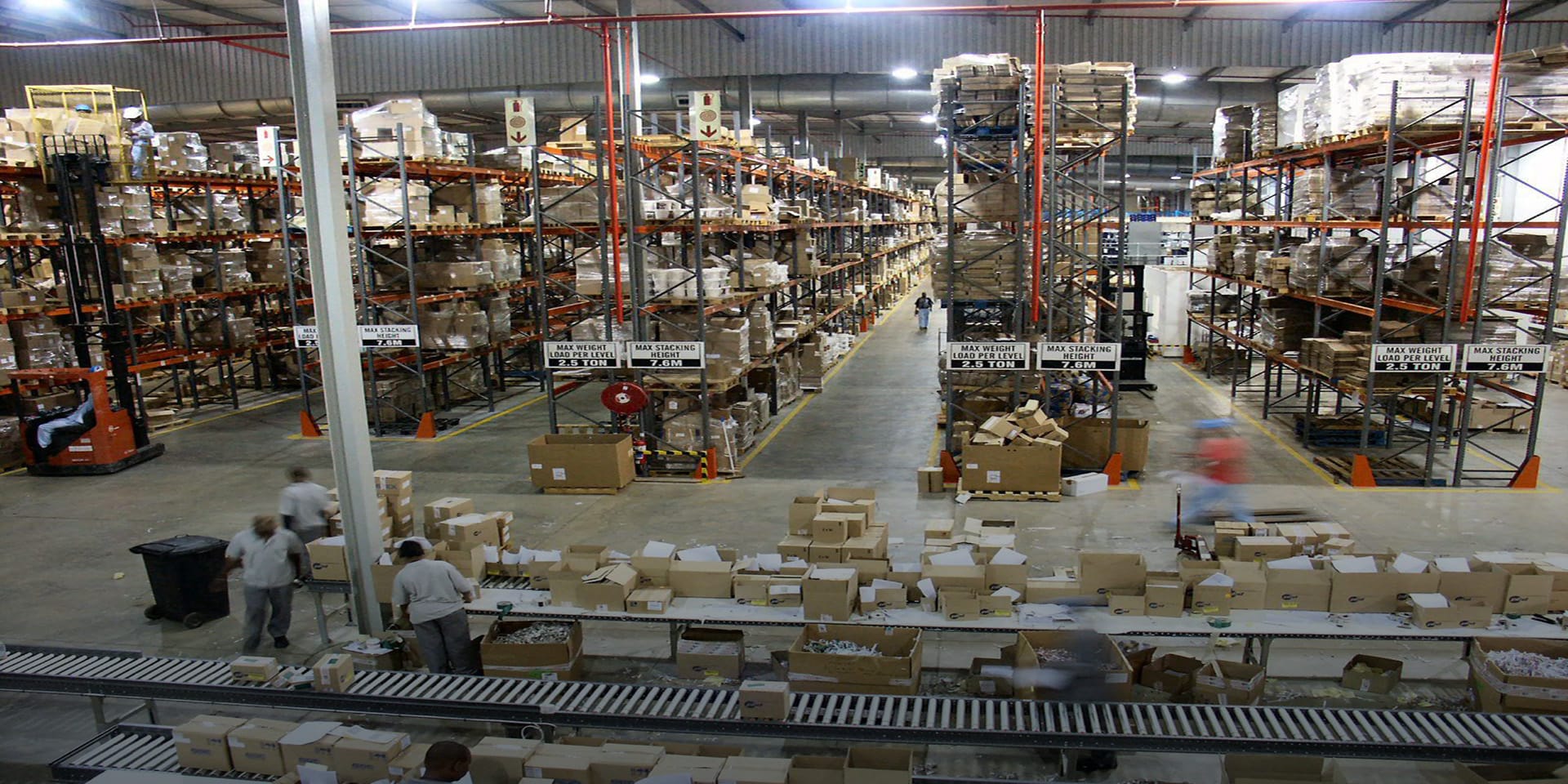 Image of a large warehouse with workers packing cardboard boxes in the foreground and several aisles of steel shelves holding boxes in the background.