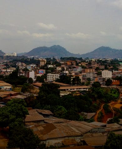 The skyline of a large town with several white, brown, and yellow buildings. Several trees are peppered throughout, and large hills can be seen in the background.