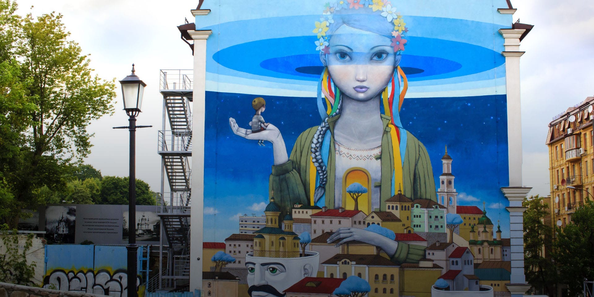A mural of a large peaceful woman towering above a city with a young boy in her hand.