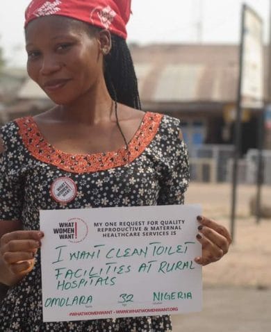 Image of a woman standing in a village. She is smiling and holding a sign that says "I want clean toilet facilities at rural hospitals."